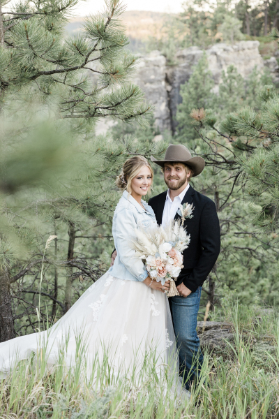 A bride and groom are surrounded by tall grass and pine trees as they pose for a wedding day picture. She wears a light blue jean jacket over her wedding dress and he wears a cowboy hat and jeans with his black sport coat.