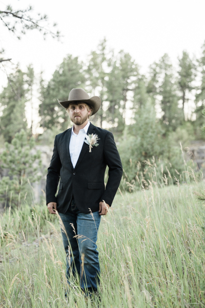 A groom poses for a wedding portrait. He stands in a grassy clearing in the woods. He sports bluejeans, a cowboy hat and a beard.