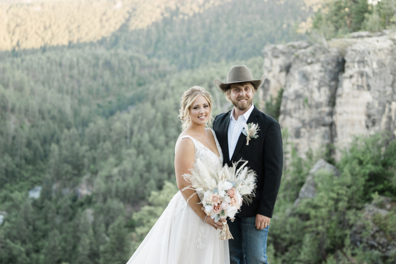 A bride and groom pose in the late afternoon sun at the top of a mountainous hillside with pine trees and a rocky cliff filling the valley behind them.