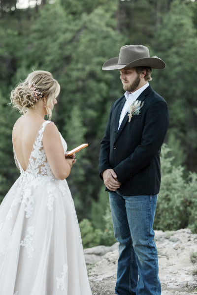 A bride reads vows from her phone as her groom stands listening with his hands folded in front of him at his belt line.