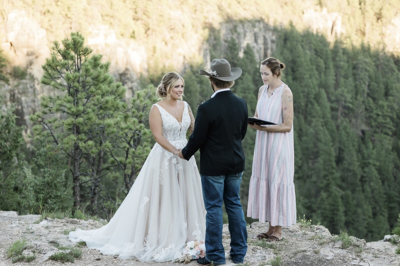 A bride and groom face each other and hold hands as they listen to their wedding officiant perform a wedding ceremony. The three of them stand near the edge of a rocky cliff in the Black Hills of South Dakota.