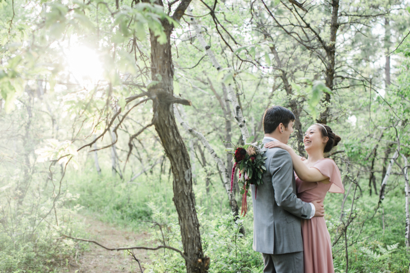 A man and woman who have just gotten married stop for a photo on a path in the woods. They are surrounded by green leafy trees and sunshine.