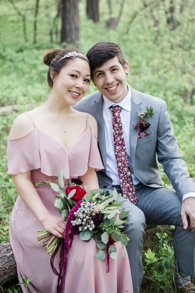 A bride in a mauve pink dress and a groom in a grey suit sit next to each other on a log in the forest. They are posing for wedding photos in the Black Hills of South Dakota.