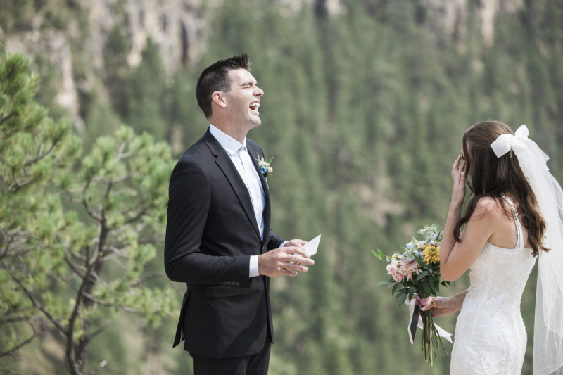 A groom reading vows to a bride. He throws his head back and his mouth open in laughter as she wipes her eyes.