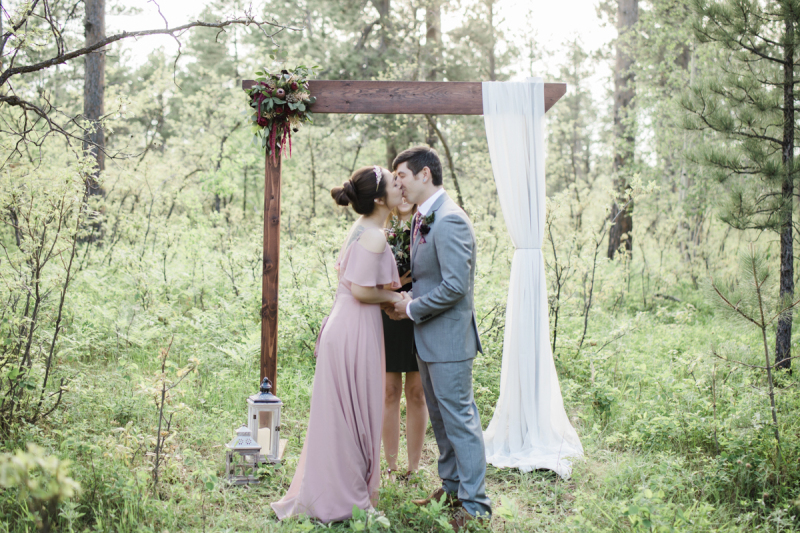 A couple kisses at the conclusion of their wedding ceremony. They are standing in front of a wooden arbor which has been placed in the woods and is surrounded by greenery.