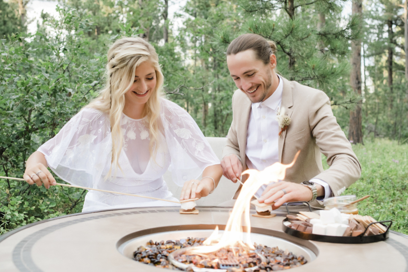 A bride and groom sit at a gas fire pit and make s\'mores on their wedding day in the Black Hills of South Dakota.