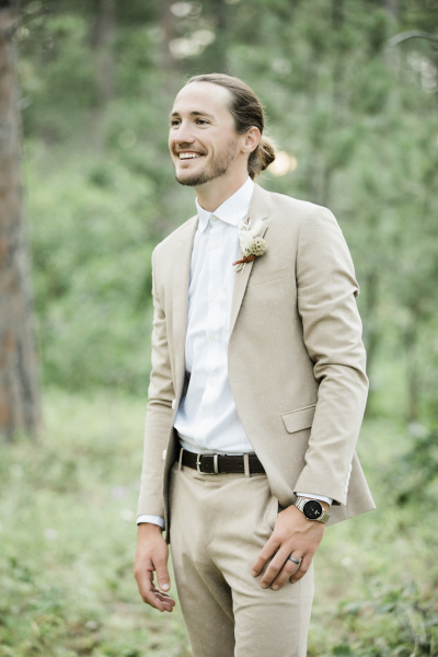 A groom in a tan suit with a white shirt stands in a forest and smiles as he looks off camera presumably as his bride approaches.