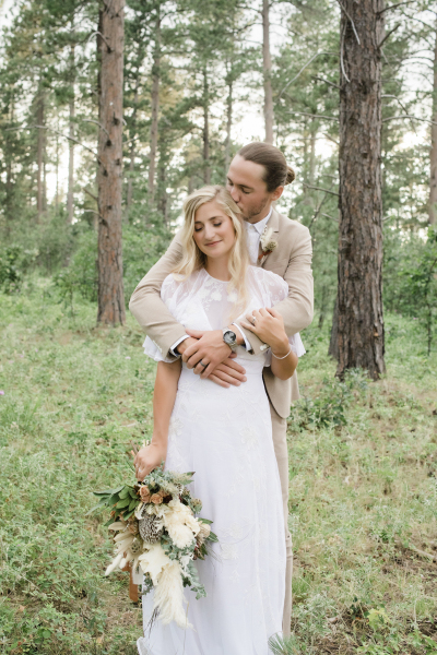 A bride and groom embrace in the middle of a pine tree forest. She stands in front of him as he reaches over her shoulders and gives her a kiss on the back of the head.