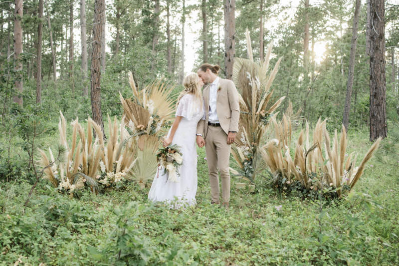 A bride and groom kiss in the woods on their wedding day. They are standing in front of a floral display made of dried palm leaves and pampas grass.