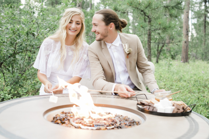 A bride and groom roast marshmallows over a gas firepit. A tray of s\'more supplies sits on the table-surround of the fire pit.
