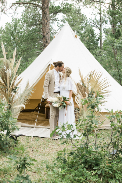 A groom and bride kiss in front of the opening of a teepee that has been erected in the woods in the Black Hills of South Dakota.