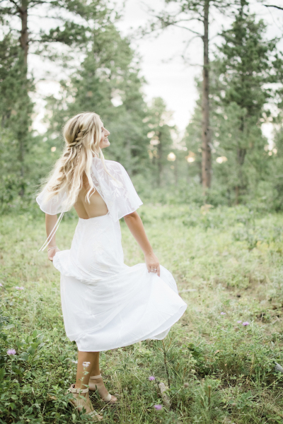 A bride is pictured in a grassy meadow in a forest. She sashays happily as she lifts and spreads the corners of her dress dramatically up off the ground in excitement.