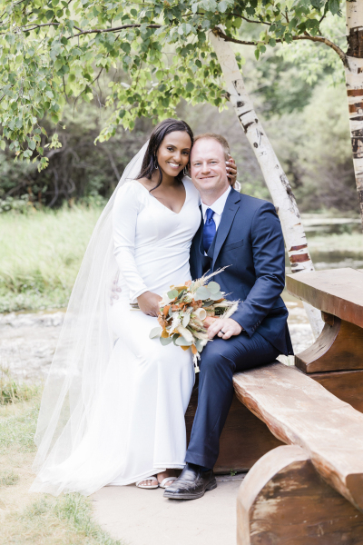 A mixed-race couple sit on a log picnic table at the edge of a stream on their wedding day in the Black Hills of South Dakota.