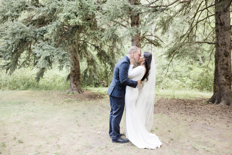 A groom and bride kiss on their wedding day. They are standing on a grassy patch underneath some pine trees in the Black Hills of South Dakota.