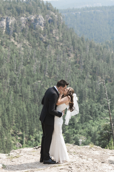 A groom and bride embrace for a kiss. They stand near the edge of a cliff in the Blacks Hills of South Dakota.