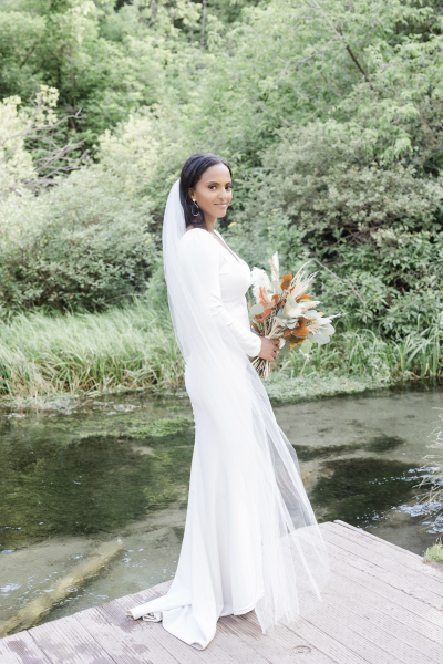 A bride poses for the camera on her wedding day. She stands at the corner of a low wooden dock that sits beside a stream. The creek beds are covered in lush vegetation and there is a log visible below the water\'s surface.