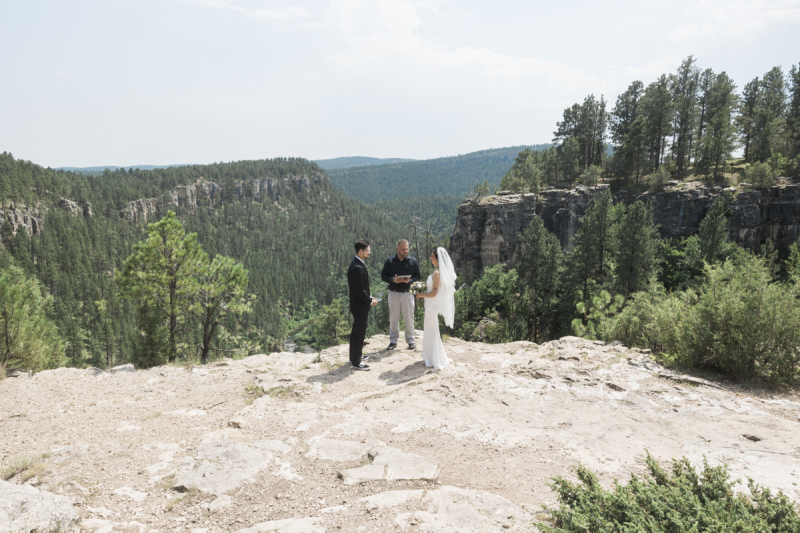 A wide shot of a groom and bride and a wedding officiant during an elopement ceremony in the Black Hills of South Dakota. The three of them are standing near the edge of a cliff overlooking a pine tree filled valley.