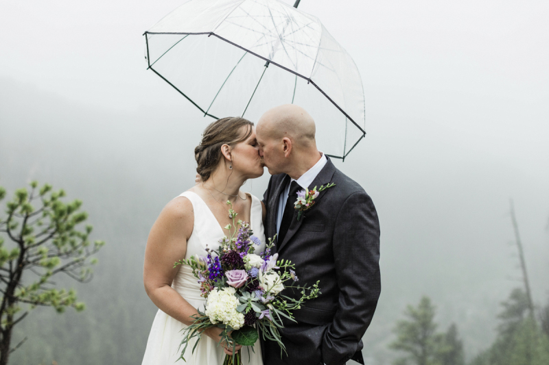 A bride and groom kiss on a foggy and rainy day underneath a clear plastic umbrella.