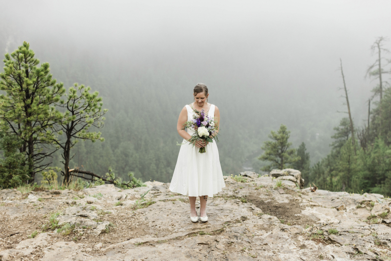 A bride stands near a rocky cliff edge in the Black Hills of South Dakota. She is holding flowers in front of her. The pine trees in the background are covered in fog.