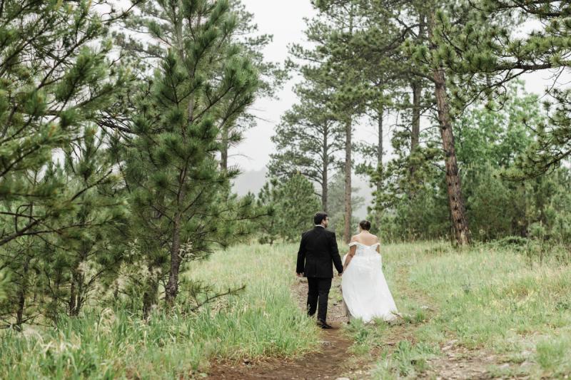A groom and bride hold hands as they walk off into the distance. They are heading down a path that leads through a clearing in the woods.