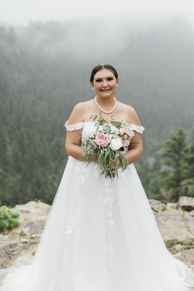 A wedding portrait of a bride in a long white wedding dress. In the background there is a fog covered pine tree forest.