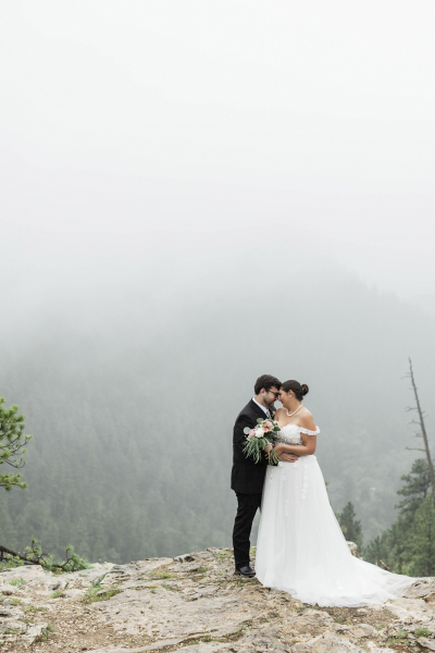 A wide shot of a groom and bride pressing their foreheads together as they pose for wedding photos at the edge of a cliff in the Black Hills of South Dakota. It is a foggy day.