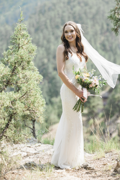 A bride poses for a full body photo of her and her wedding dress. She holds a bouquet of flowers as a breeze catches her veil and pushes it to the side.