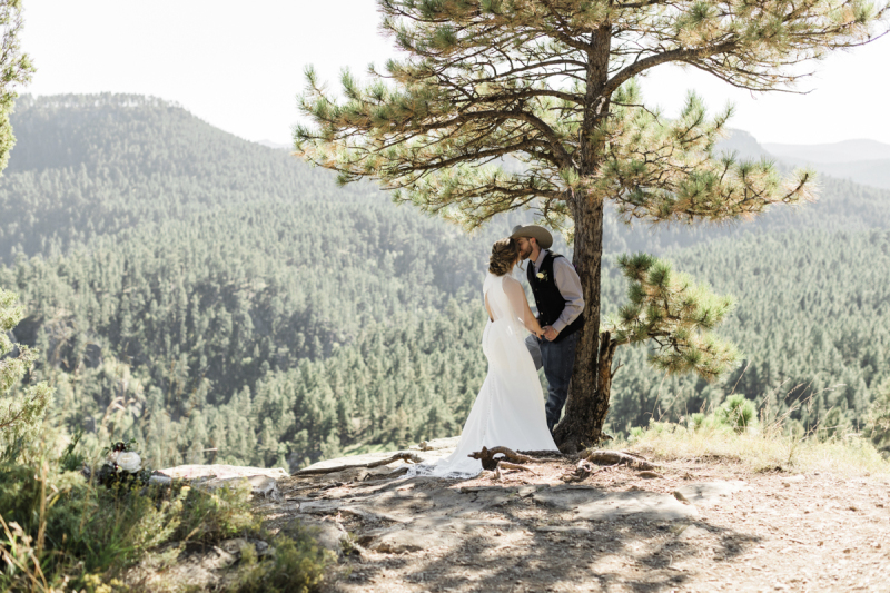 A bride and groom kiss underneath a pine tree that is growing at the top of a rocky hill. He wears blue jeans and a cowboy hat and leans against the tree.