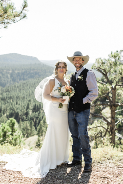 A bride and groom stand together and pose for a wedding portrait following their wedding ceremony in the Black Hills of South Dakota. She wears a traditional white dress and he wears jeans, a vest and a cowboy hat.