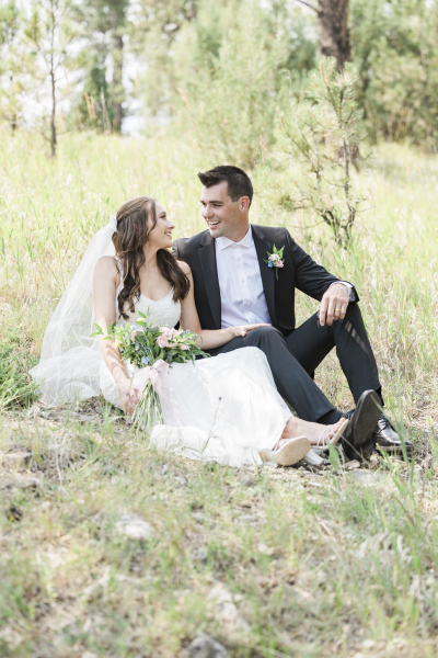 A bride and groom sit on the ground and look at each other lovingly. They are posing for wedding photos in a grassy meadow in the Black Hills of South Dakota.