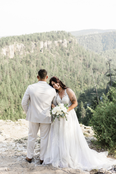A groom has his back to the camera as he looks out over a pine tree covered valley. His bride is leaning against his side while facing the camera and smiling.