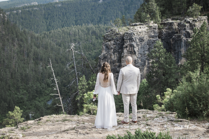 A bride and groom hold hands and face away from the camera as they look out over a pine tree covered valley in the Black Hills of South Dakota.