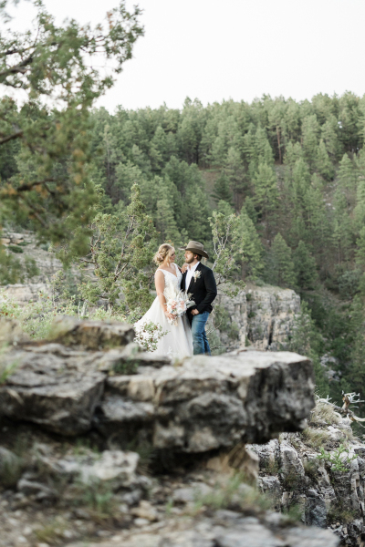 A rocky cliff-side artfully frames a couple on their wedding day. The bride and groom are smiling as they face each other.