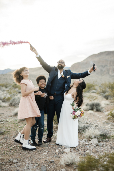 A wedding celebration takes place in the desert outside of Las Vegas, Nevada. A groom and bride are joined by some children. The group all look to the sky as the groom holds a colorful smoke bomb in each hand that emit purple and white smoke.