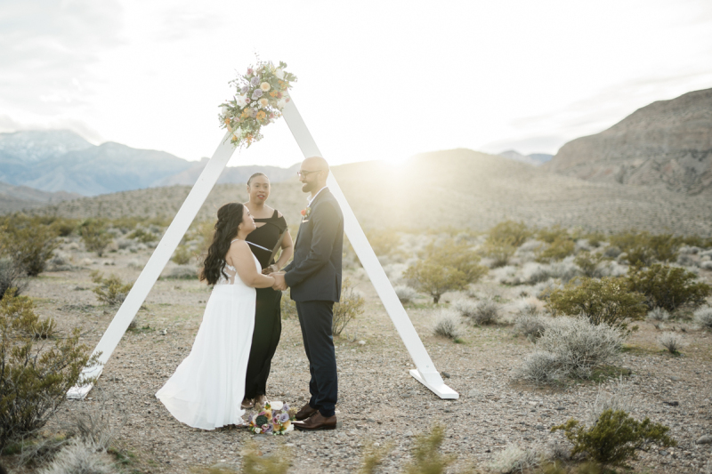 A bride and groom stand facing each other holding hands. They are smiling and listening to the words of the officiant of their desert wedding. The late afternoon sun shines through the simple, white, triangular arbor which decorates the desert west of Las Vegas.