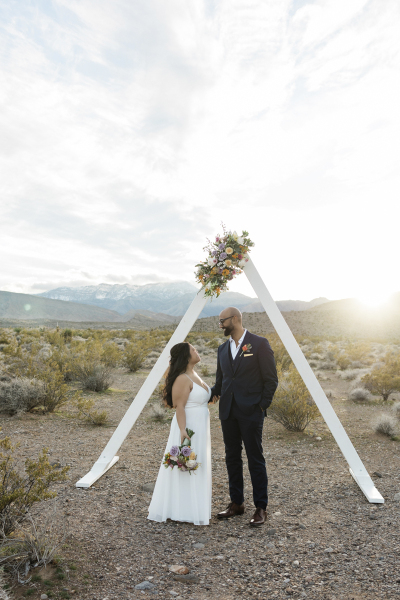 A bride and groom stand beneath a large triangular arbor placed in a desert landscape. The sun is setting behind them. The white arbor is decorated with a large bouquet of flowers at the pinnacle and the couple looks at each other lovingly.
