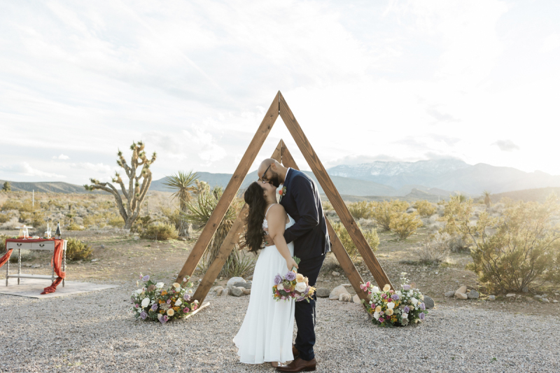 A bride and groom kiss underneath a wooden double-triangle shaped arbor. They are being wed in the Nevada desert outside Las Vegas. Joshua trees, Creosote bushes, Yucca plants and a mini reception table fill the scene as the late afternoon sun sets off-camera to the right.