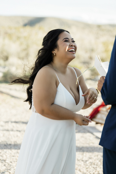 A bride laughs as her groom reads his vows to her. The photograph is a medium shot of the bride with the groom's torso and left arm and hand visible on the right of the frame. The desert landscape behind the bride is brightly lit by the sun.