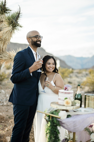 A groom and bride share a glass of Champagne to celebrate their desert wedding. They are standing underneath a Joshua tree and in front of a mini reception table with a cake, an ice bucket and a Champagne bottle.