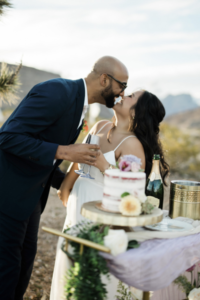 A groom leans over to kiss his bride. They are holding Champagne glasses and toasting their outdoor desert wedding. A cake, ice bucket and Champagne bottle sit on the mini reception table visible in the foreground.