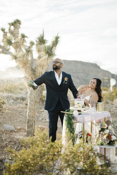 A groom and bride spray Champagne from a bottle to celebrate their desert wedding. They are standing underneath a Joshua tree. A wedding arbor is seen in the background, and mountains are visible in the distance. 
