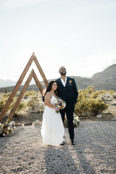 A bride and groom stand side-by-side at a triangular wooden arbor decorated with flowers. Their wedding is taking place in the Mojave Desert on the western edge of Las Vegas.