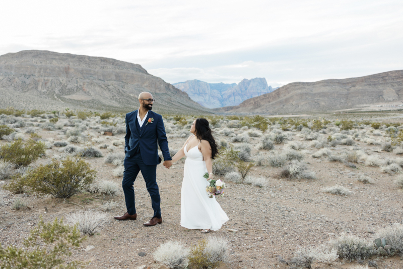 A groom and bride walk hand-in-hand through the desert outside of Las Vegas, Nevada on their wedding day. In the background the mountains of the Red Rock Canyon National Conservation Area are seen.