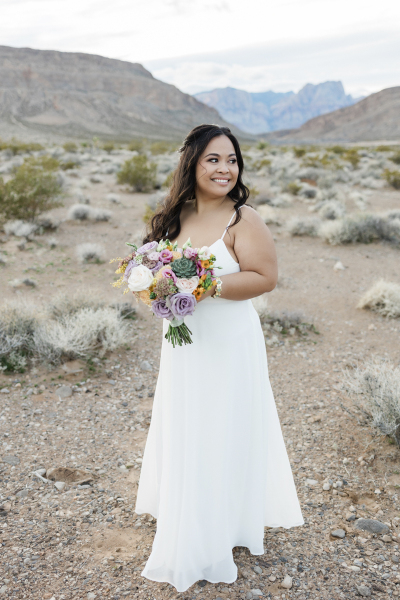 A bride in a simple floor length wedding dress holds a bouquet of flowers while posing for a photo in the Nevada desert outside of Las Vegas. She looks off camera to the right. In the background, the mountains of the Red Rock Canyon National Conservation Area rise majestically.