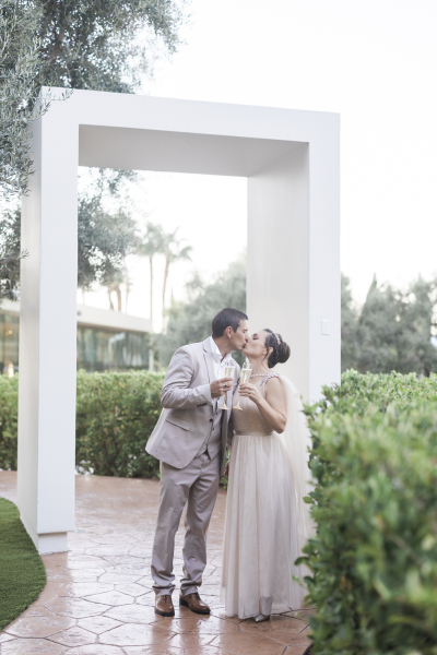 A groom and bride kiss and hold Champagne flutes underneath a large rectangular arch spanning a garden sidewalk.