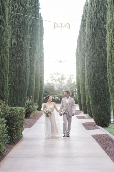 A bride and groom hold hands as they walk towards the camera between two rows of cypress trees.