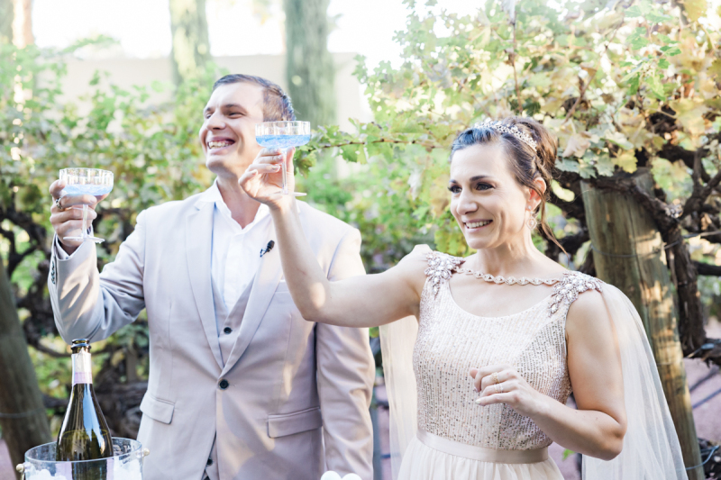 A groom and bride smile and raise Champagne glasses as they stand in front of grape vines.