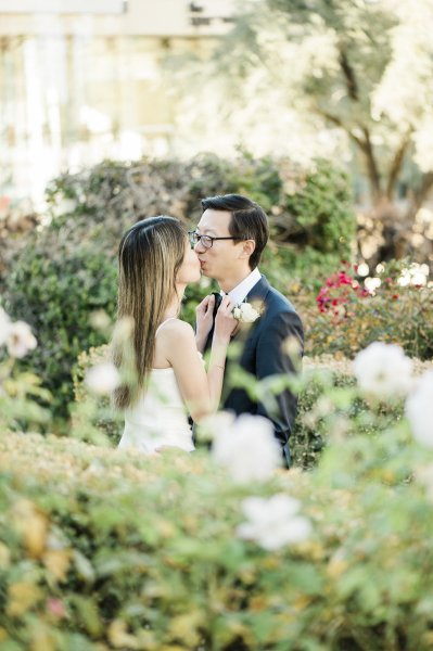 A bride and groom kissing in a rose garden as she ties his bowtie.
