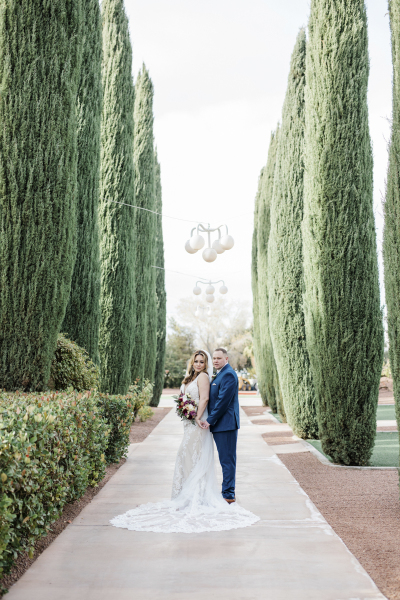 A bride and groom pose for wedding photos on a sidewalk that splits two rows of cypress trees.