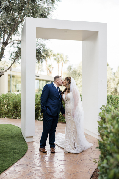 A couple kisses underneath a large rectangular archway that spans across a garden sidewalk.